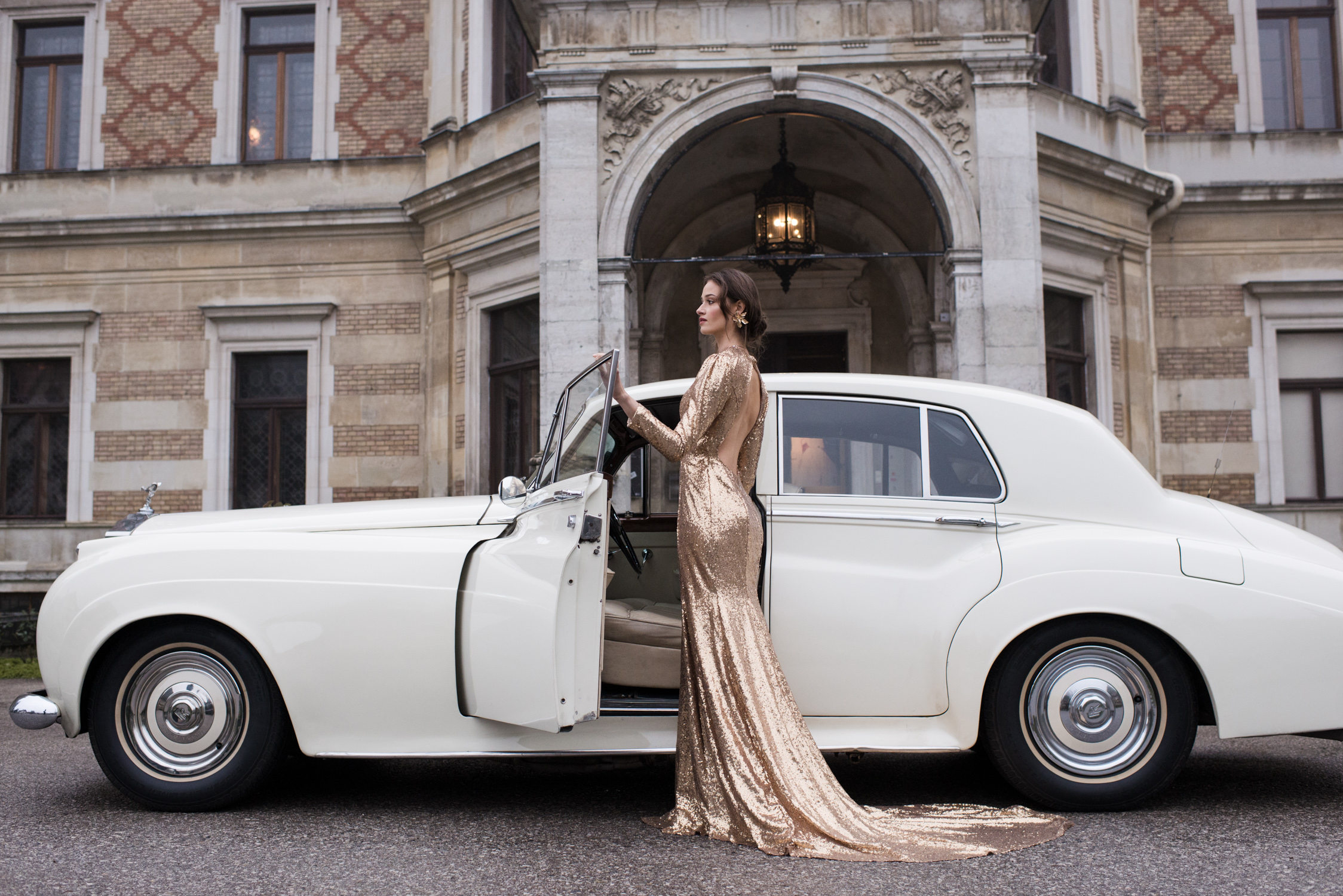 woman in golden paillette dress with her rolls royce car in front of her villa